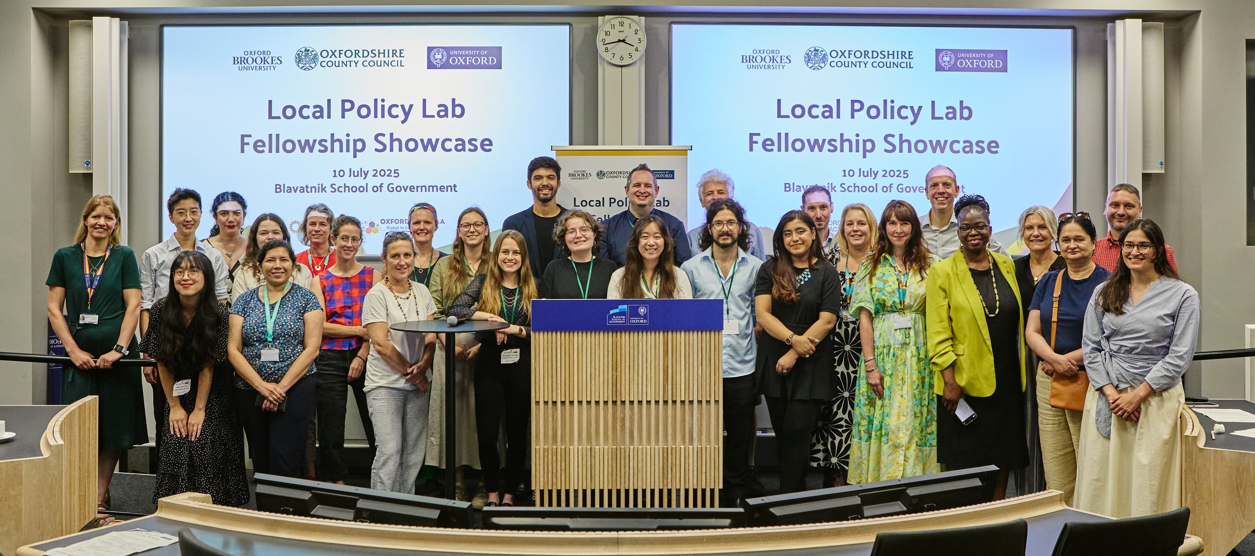 A group of more than 20 postgraduate researchers, academics and policymakers stand in front of a presentation entitled 'Local Policy Lab Fellowship Showcase'