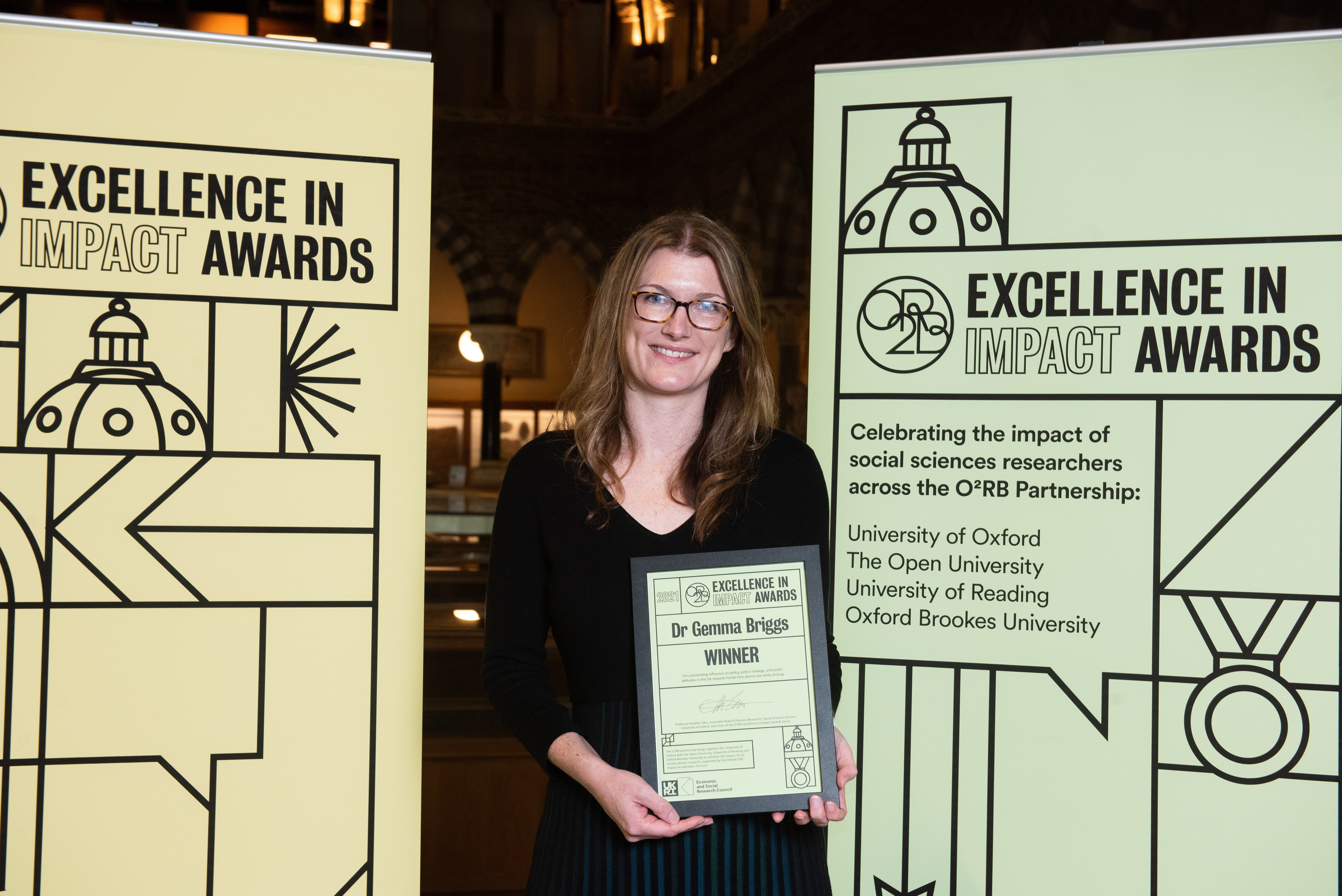 Gemma Briggs smiles as she holds her Impact Awards winners certificate in front of the O2RB Excellence in Impact Awards event branding