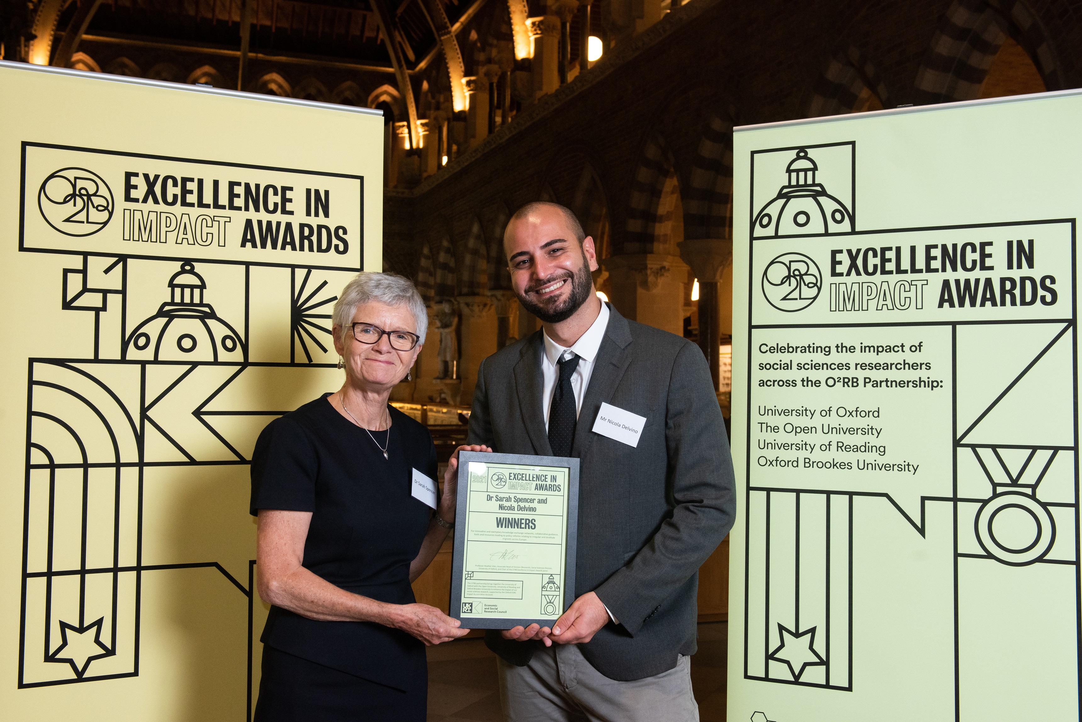 Dr Sarah Spencer and Mr Nicola Delvino smile as they hold their Impact Awards winners certificate, in front of the O2RB Excellence in Impact Awards event branding