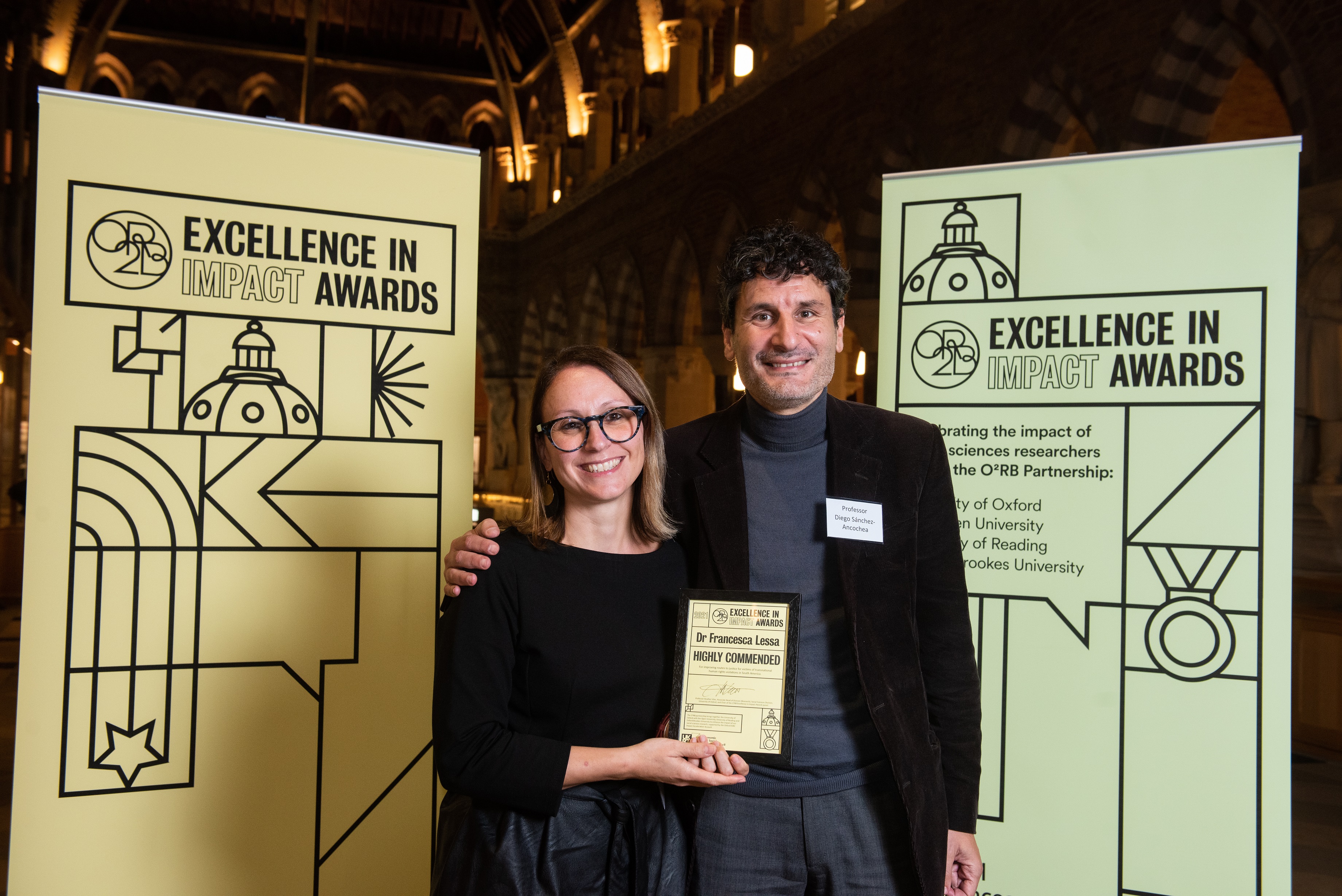 Dr Francesca Lessa holds her Excellence in Impact Awards Highly Commended certificate, standing next to Prof Diego Sánchez-Ancochea and flanked by the event branding