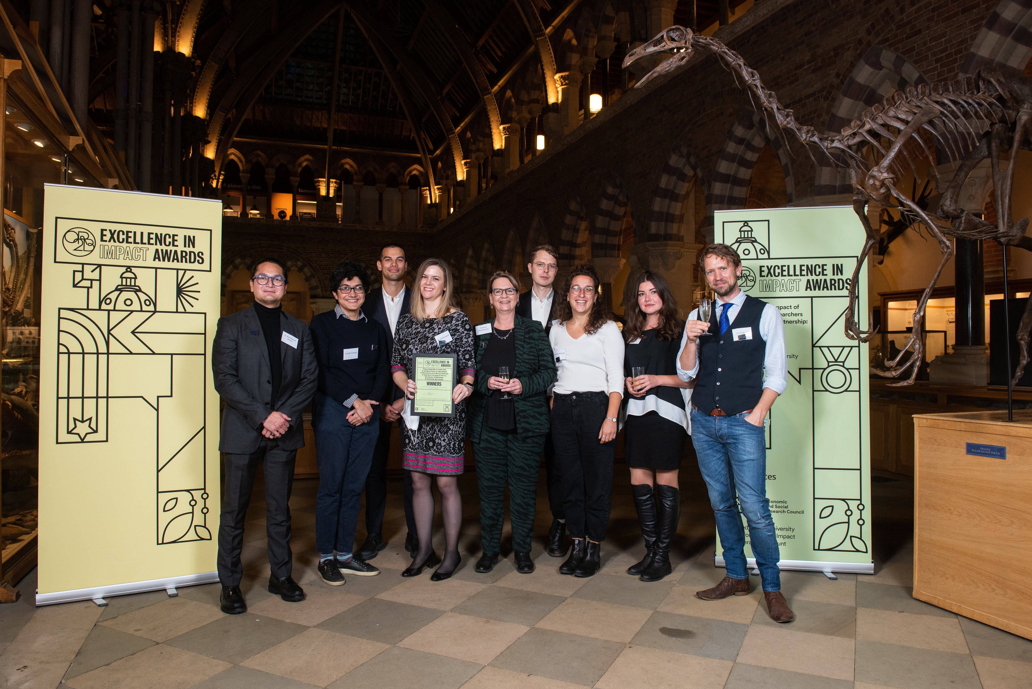 Professor Melinda Mills, Dr Jennifer Beam Dowd, and colleagues from the LCDS team smile as they hold their Impact Awards winners certificate, in front of the O2RB Excellence in Impact Awards event branding