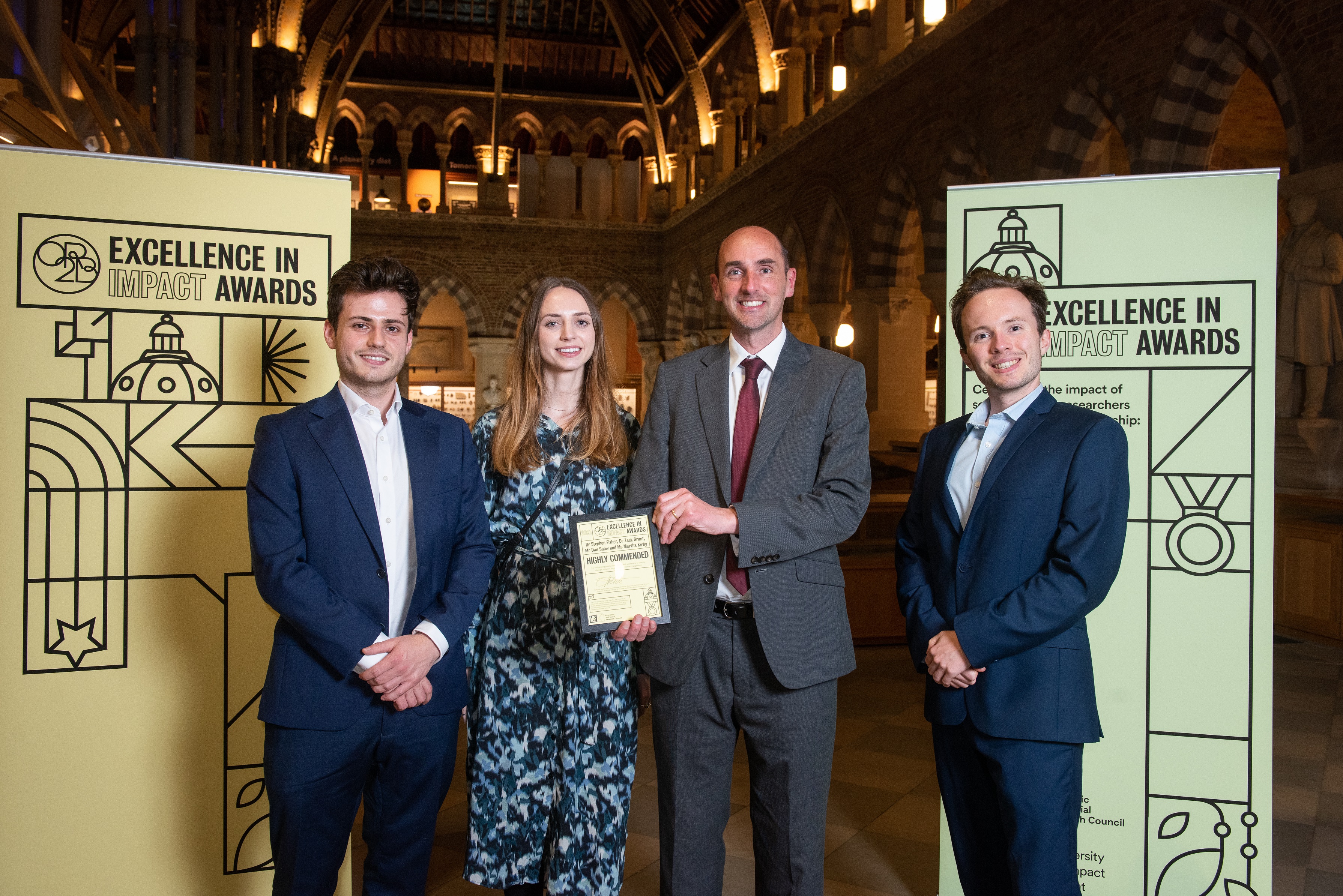 Mr Dan Snow, Ms Martha Kirby, Dr Stephen Fisher and Dr Zack Grant smile as they hold their Highly Commended certificate, flanked by impact awards branding at the Museum of Natural History
