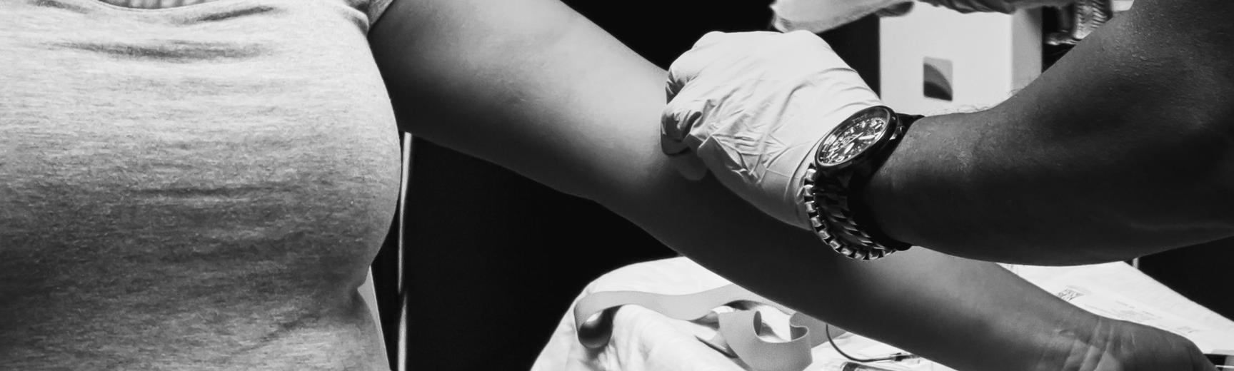 A black and white image of a medical professional, who is wearing gloves, preparing a patient's arm for an injection