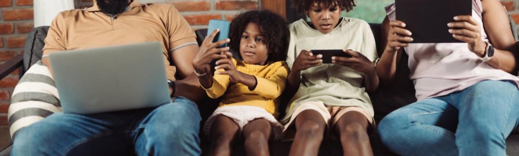 A family of two adults and two children sitting on a sofa, each looking at their own digital devices.