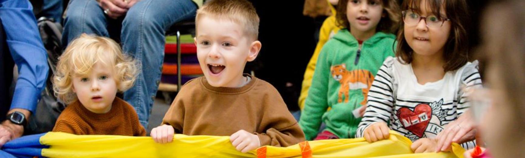 Several young smiling children waving a large, brightly coloured cloth between them.