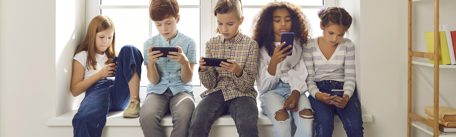 Group of children of different nationalities sit in a row on the windowsill and with lack of emotion play online games or read social networks on mobile phones