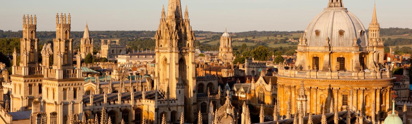 An image of the Radcliffe Camera and All Soul's College taken from rooftop level. The buildings are lit up by golden hour sunlight, against a blue sky. 