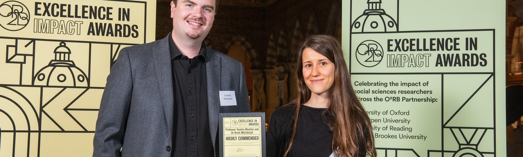 Professor Sandra Wachter and Dr Brent Mittelstadt hold their Highly Commended impact award, flanked by event branding at the awards reception