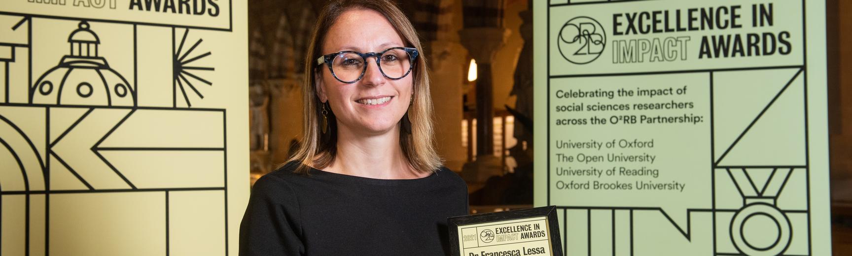 Dr Francesca Lessa holds her Excellence in Impact Awards Highly Commended certificate at the Awards reception evening, flanked by pull-up banners of the awards branding. In the background are the arches of the Museum of Natural History. The banners read: 