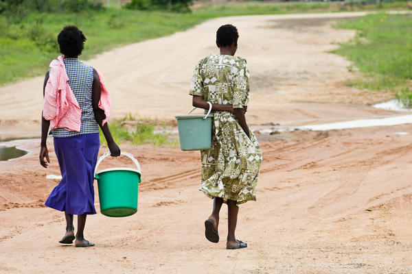 The view from behind of two women from an African village carrying empty buckets to collect water. The ground is mostly arid and dry, and there is grass in the distance.