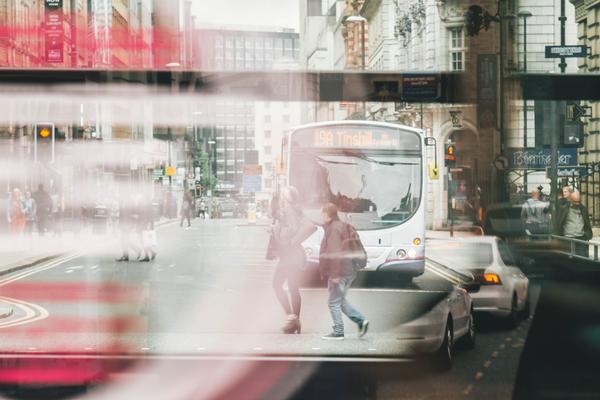 Photo of a bus window reflecting people walking and a bus and cars driving through a street. The image is slightly blurred and gives the impression of movement.
