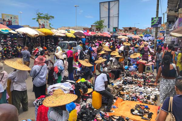 A busy market scene where people are buying and holding clothes and shoes. Many people are wearing sunhats and the market stalls are shaded by sun umbrellas.