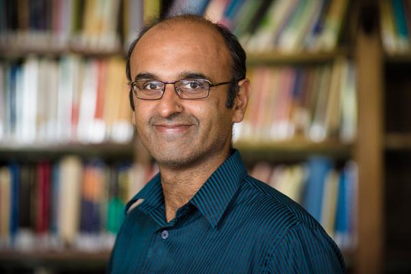 Portrait photo of Professor Yadvinder Malhi, smiling against a blurred background of shelves full of books