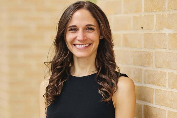 Dr Hawbaker has long wavy brown hair is wearing a black sleeveless top, and is smiling whilst standing by a brick wall.