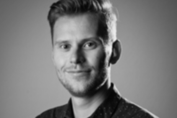 A black-and-white portrait of Dr Brindle with short hair and a trimmed beard, smiling slightly. He is wearing a dark, button-up shirt with a small dotted pattern