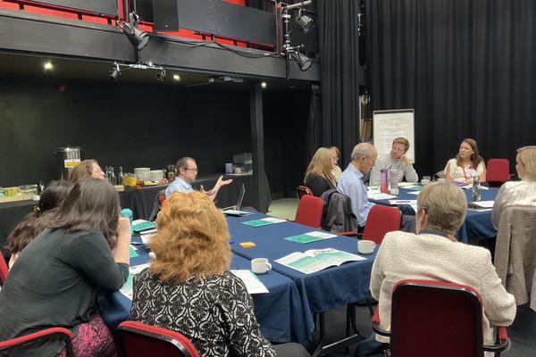 Participants seated around conference tables engage in a workshop discussion, with one individual presenting to the group.