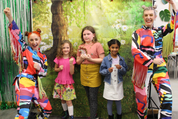 Two adults, dressed as mutli-coloured, cheering butterflies, and three smiling children, pose  as “superpillars” in front of a forest backdrop.