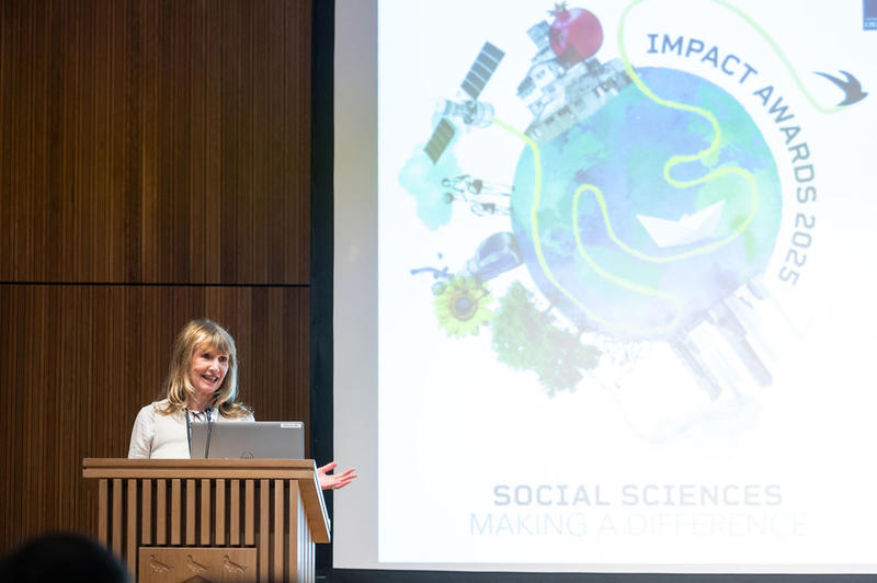 Dr Jen Gold presents the Developing Impact category Prof Tina Miller speaks and gestures from the lectern. Behind her is a screen with the Impact Awards branding.