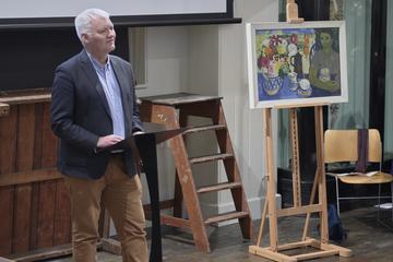 Associate Professor Ian Thompson is standing in front of a ladder and some art. He is wearing brown trousers, blue shirt and navy blazer and presenting to the room. 