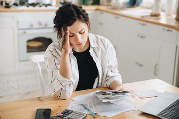 A woman with dark hair sits at a desk, wearing a black tshirt and white overshirt. She looks stressed, resting her head on one hand, while looking at piles of receipts next to a calculator. Credit: sheremetio - stock.adobe.com