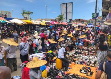 A busy market scene where people are buying and holding clothes and shoes. Many people are wearing sunhats and the market stalls are shaded by sun umbrellas.