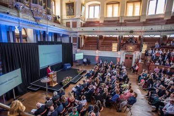 An audience in the Sheldonian Theatre. They are facing a stage, where the Vice-Chancellor is speaking at a podium, wearing a red suit. 