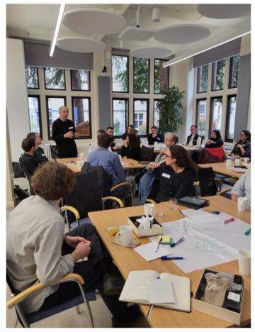 A male academic addresses a room full of researchers who listening to him attentively, and seated around tables that are covered in pens and paper, brainstorming activities, and lunch