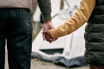 Taken from behind as an adult holds a child's hand. Together they face an out-of-focus tent in the background