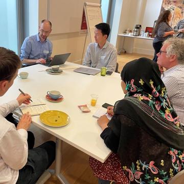 Common Ground seminar 3 Researchers seated around a table are in conversation; one is typing, two others are taking notes by hand. In the background is a flipchart and more people in conversation