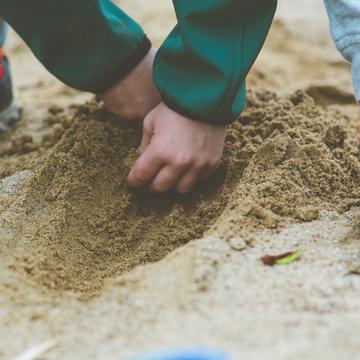 Shows a child's hands and feet as they crouch to dig a hole in the sand