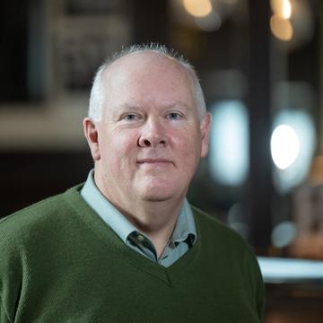 Professor Timothy Power has short white hair and is wearing a green jumper and light blue shirt