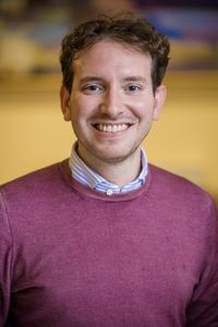 Dr Francesco Rampazzo Dr Rampazzo has short brown curly hair. He is wearing a dark pink jumper and smiling widely at the camera.