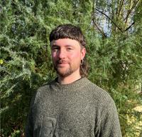 Dr Jonathon Turnbull Dr Turnbull has brown hair styled in a mullet. He is wearing a green knitted jumper and standing in front of some foliage.