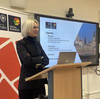 Professor Louise Slate stands at a lecture podium in front of Social Sciences Divisional branding and a slide of the agenda for the Democracy seminar