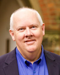 A white man with grey hair stands against a blurred background. He wears a bright blue shirt and dark blue jacket.