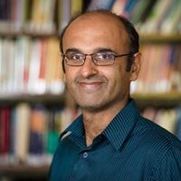 Portrait photo of Professor Yadvinder Malhi, smiling against a blurred background of shelves full of books