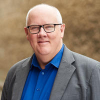Tim Power wears a blue shirt and grey jacket. He is white and has white hair, he wears glasses with black rims. He is standing outside with a brick wall behind him, which is blurred. He is smiling.