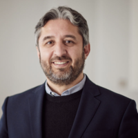 Dr Adam Mahdi Dr Mahdi has short grey and dark hair and a short beard. He is wearing a shirt, navy jumper and blazer.