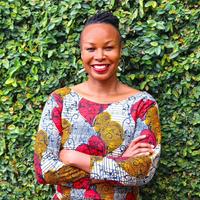 Dr Helidah Ogude-Chambert Dr Ogude-Chambert has dark braided hair and wearing a white top with patterns of bright red and yellow shapes. She is smiling and standing with her arms folded in front of some foliage.