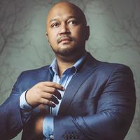 Eusebius McKaiser stands in a blue suit and shirt against a grey wall. His arms are raised