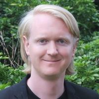 Dr Laurence Wainwright Dr Wainwright has long blonde hair and is wearing a black t-shirt and blazer. He is standing in front of foliage and smiling.