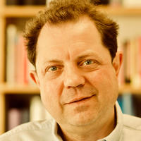 Professor Paul Irwin Crookes stands against a bookshelt. He is wearing a white open collar shirt.