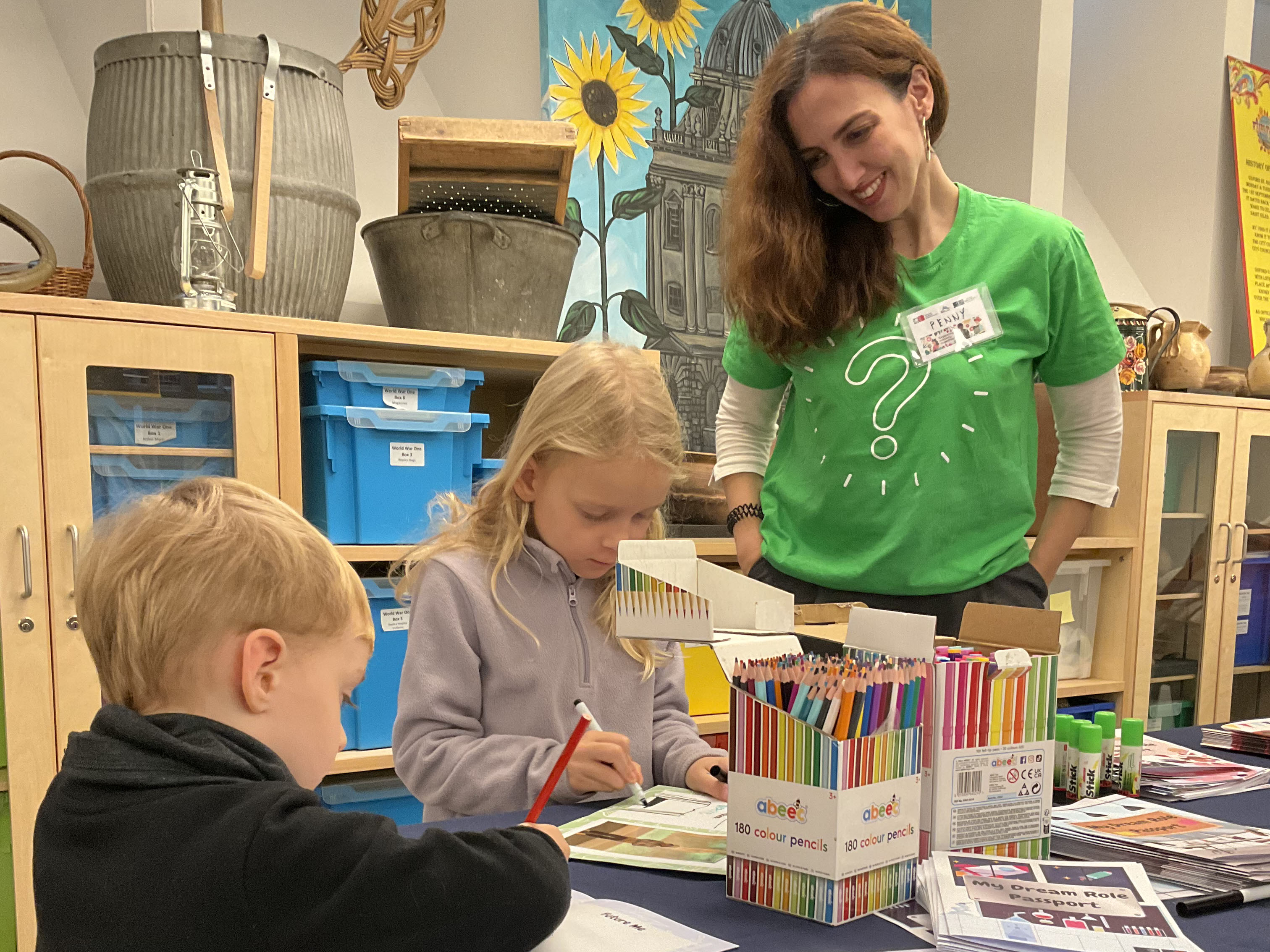 Two children using colour pencils with an adult smiling and supervising