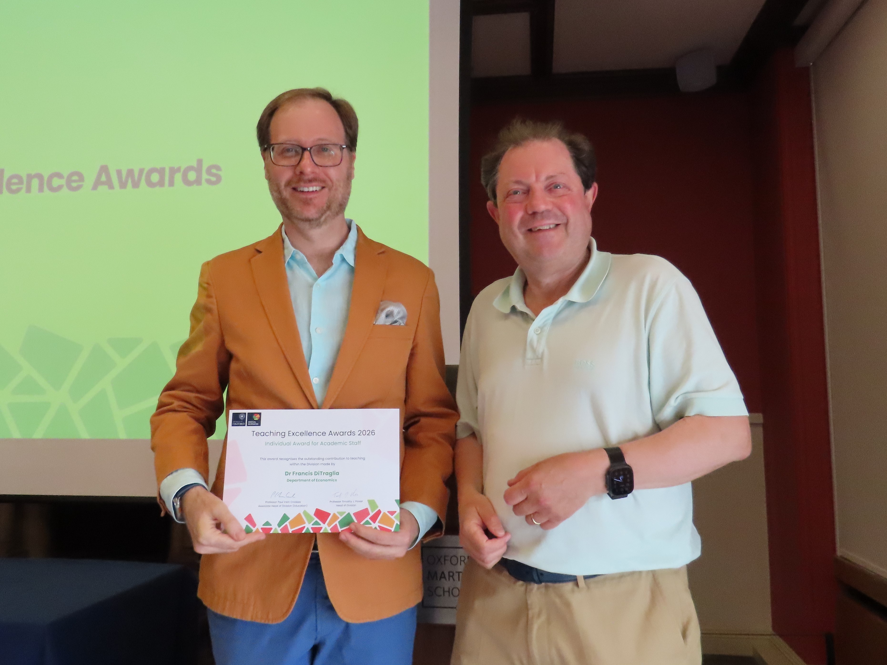 Professor Francis DiTraglia, wearing an orange blazer and holding a certificate, stands and smiles next to Professor Paul Irwin Crookes
