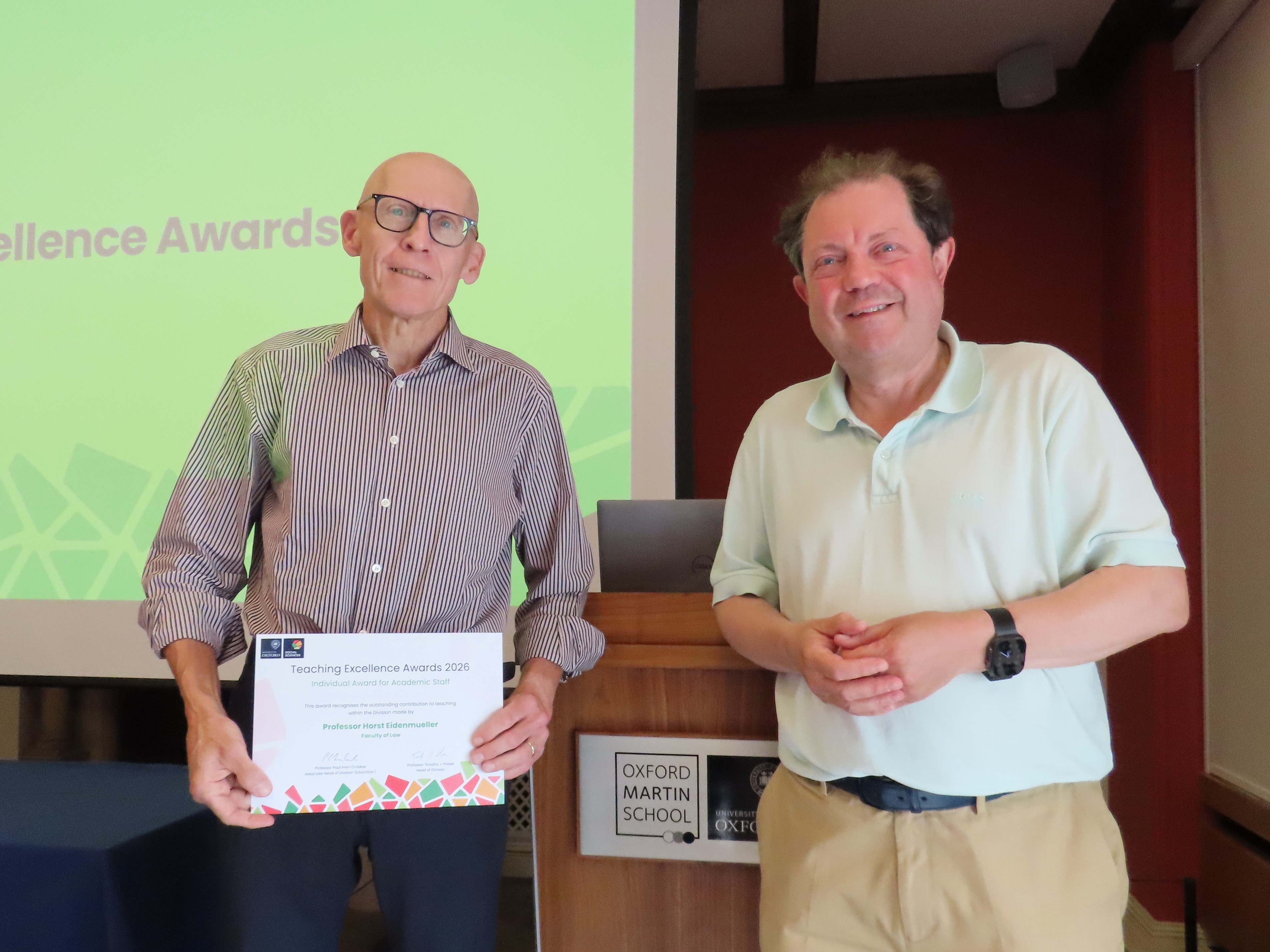 Professor Horst Eidenmuller, wearing a light checked shirt, standing on stage and holding a certificate next to Professor Paul Irwin Crookes