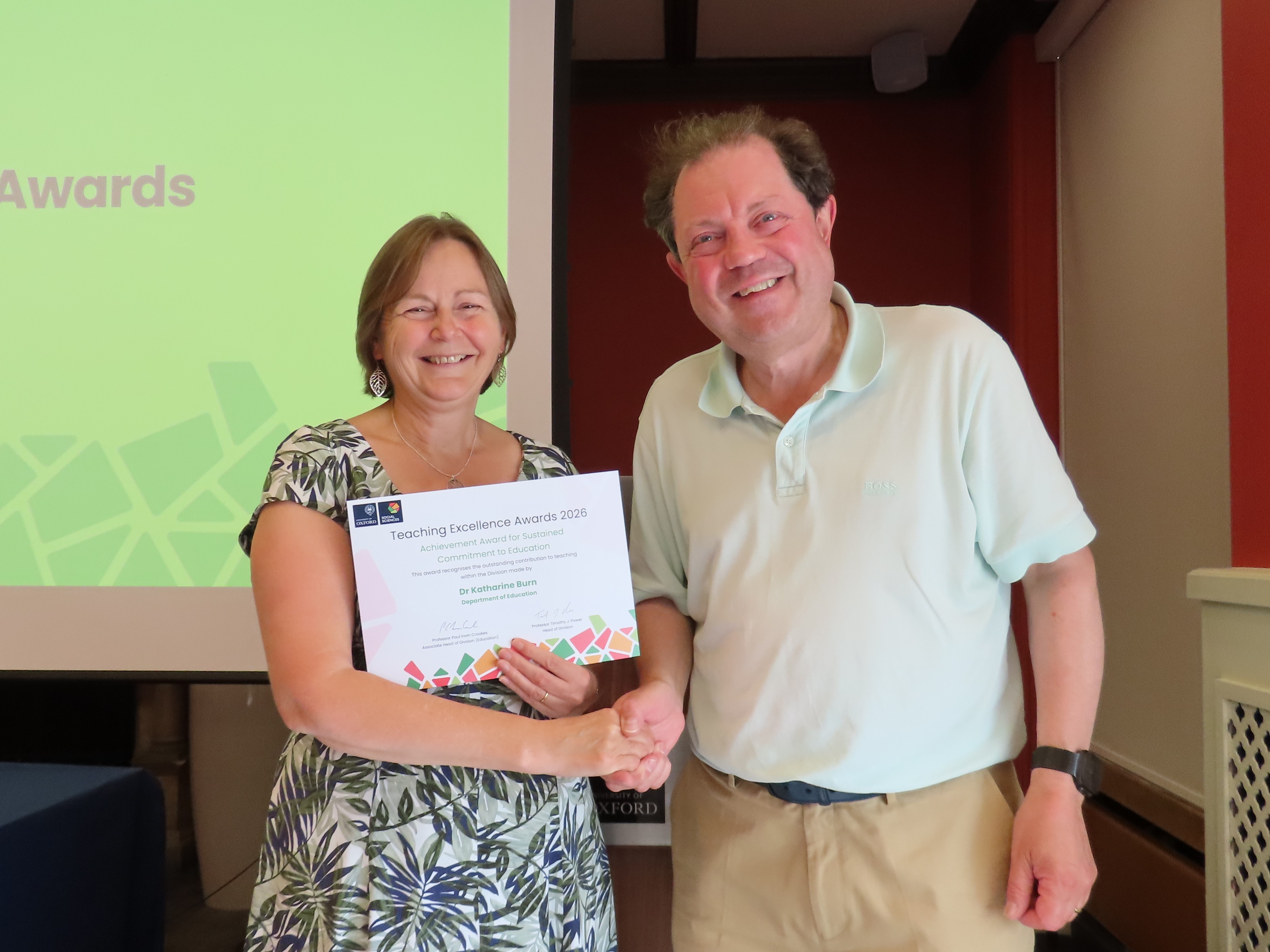 Dr Katharine Burn, wearing a light summer dress with a pattern of leaves, shakes hands with Professor Paul Irwin Crookes, holding a certificate