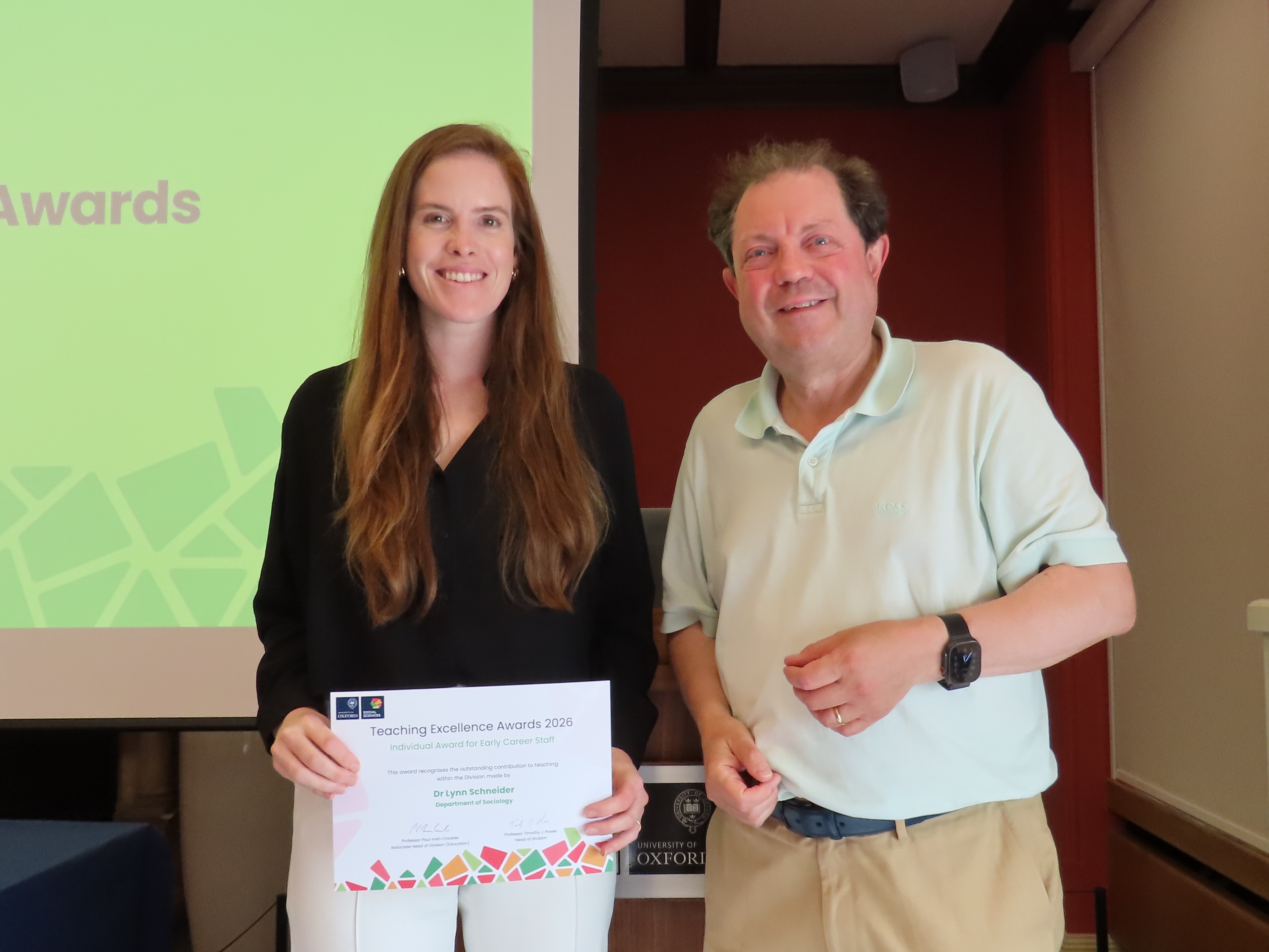 Dr Lynn Schneider, wearing a black blouse and white trousers, holding a certificate next to Professor Paul Irwin Crookes