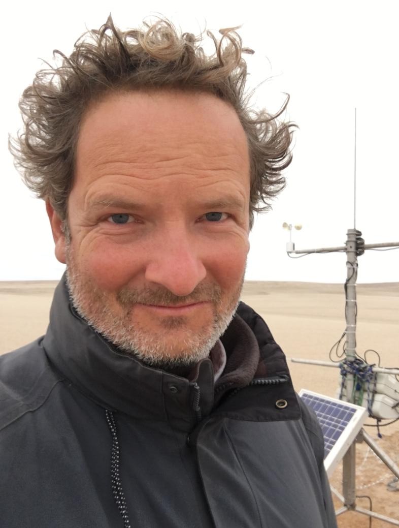 Giles Wiggs has short curly brown hair. He is wearing a black jacket and standing outside