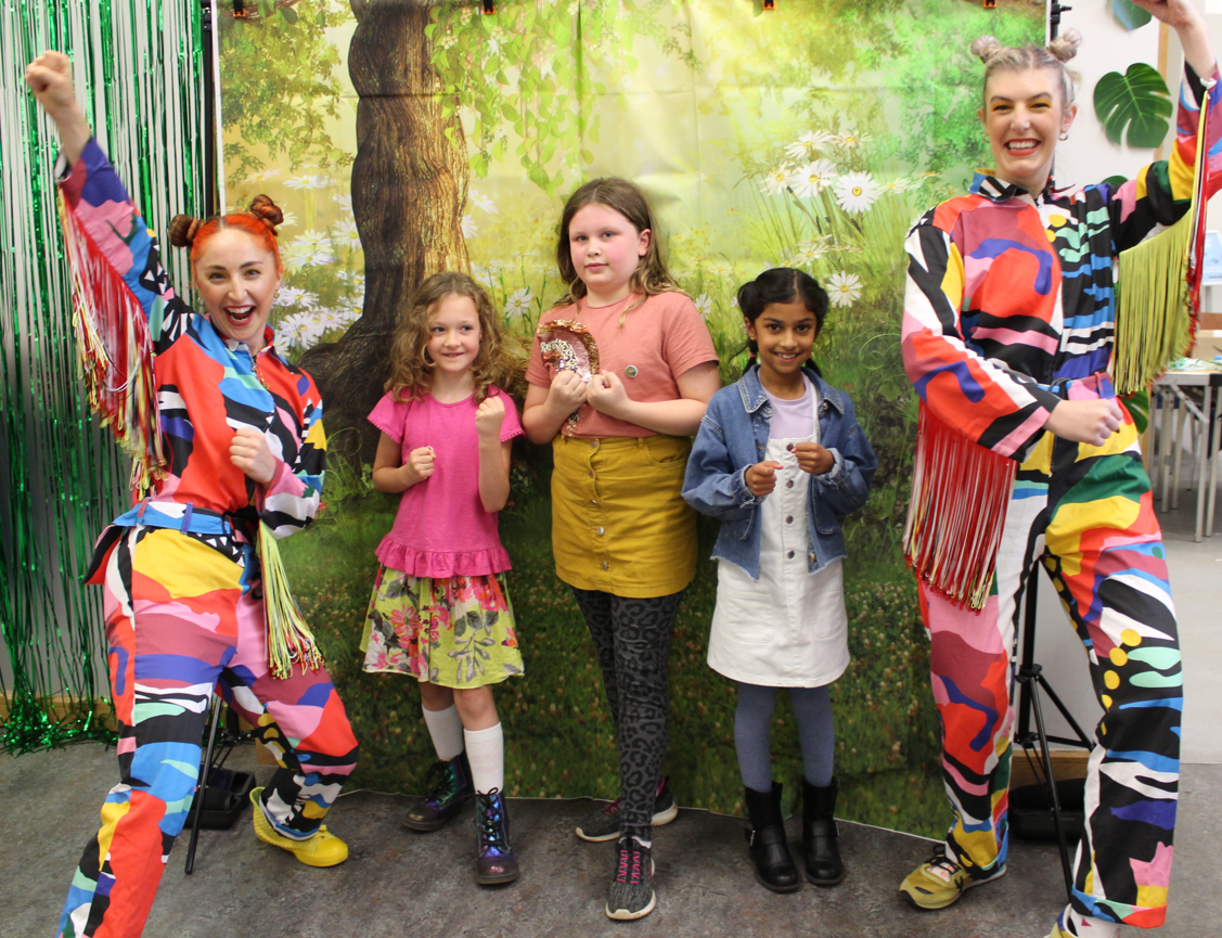 Two adults, dressed as mutli-coloured, cheering butterflies, and three smiling children, pose  as "superpillars" in front of a forest backdrop.