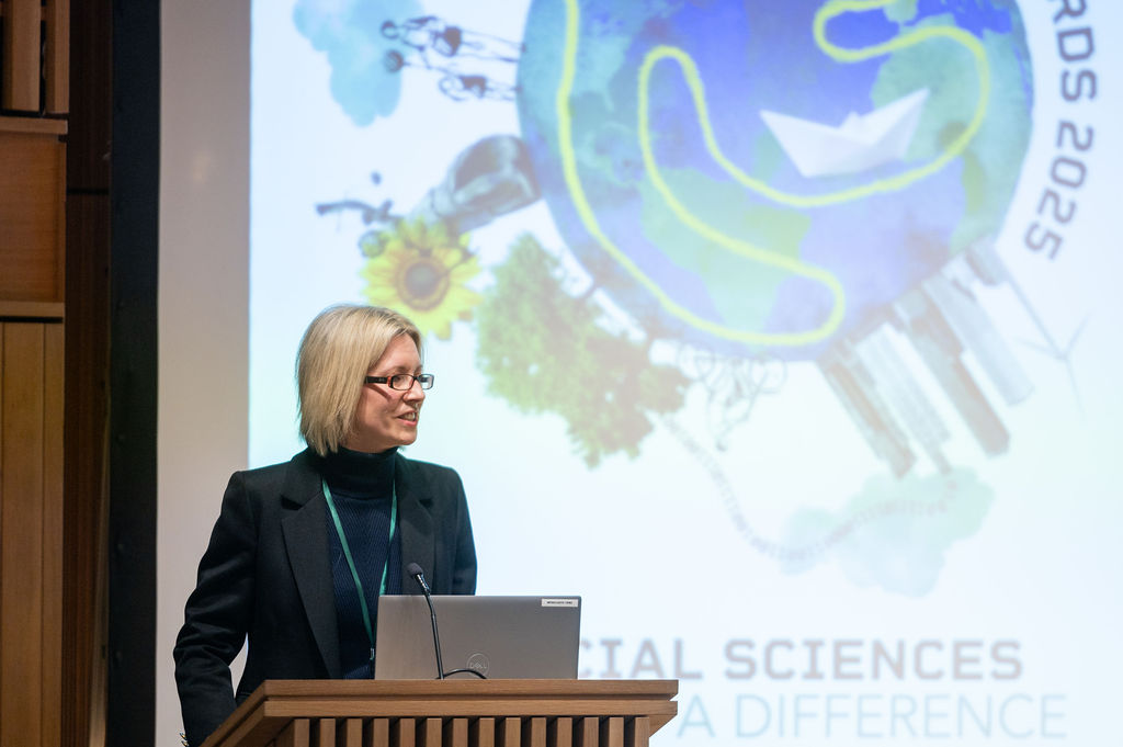 Prof Louise Slater speaks from a lectern in front of a screen with the event branding